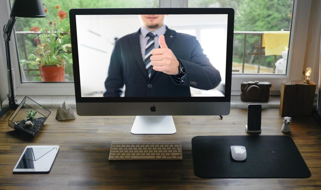 A computer monitor displaying a person in a suit giving a thumbs-up during a remote meeting.