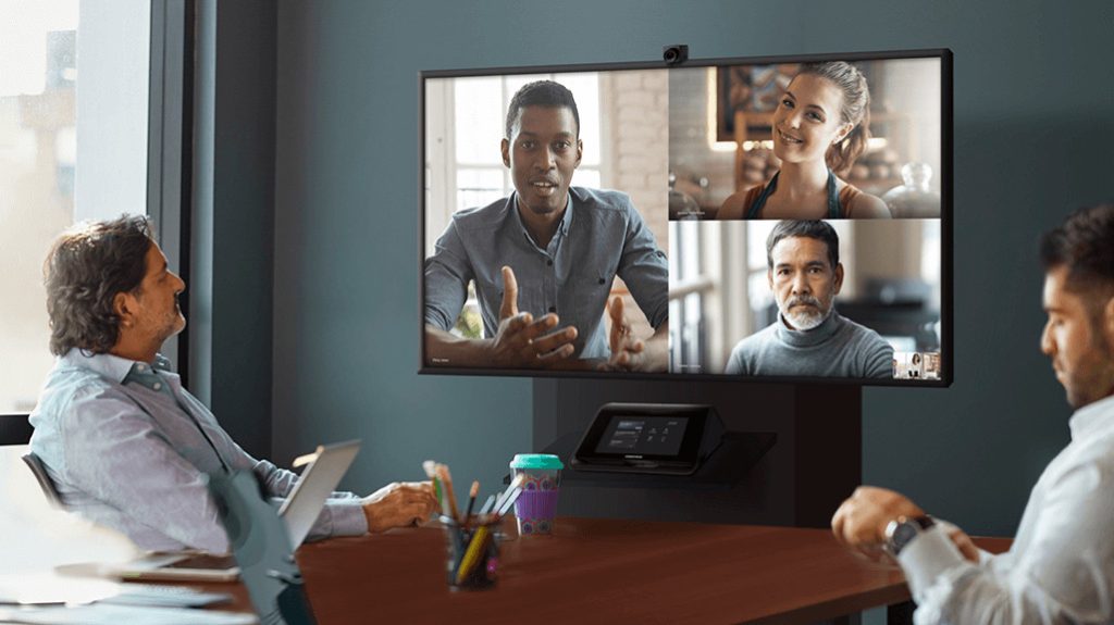 A woman and three men participate in a video conference using a large screen display.